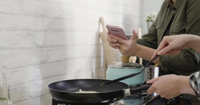 two unrecognized young asian women cooking healthy snack in kitchen. female hands using frying pan and turn pancake on stove by spatula. friend using mobile phone taking photo to record down step.