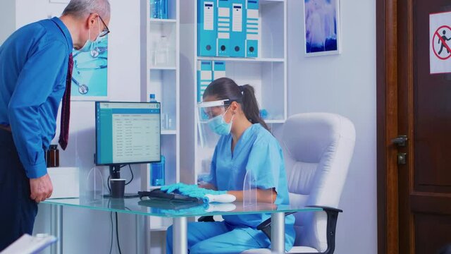 Nurse with visor against coronavirus at hospital reception greeting senior patient. Nurse and disabled woman in wheelchair passing by through waiting area. Health care system during covid-19 global