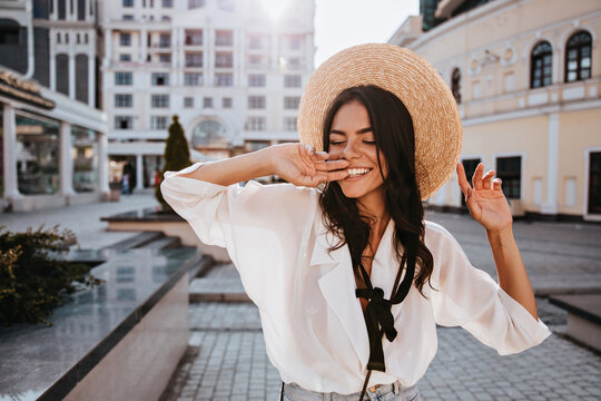Blissful woman in summer hat posing in city. Outdoor photo of blithesome brunette girl in white blouse walking around town.