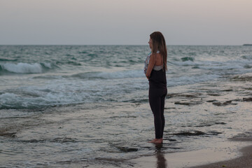 Woman meditating at the beach while standing with her feet in the water, facing the sea. Wide angle shot at sunrise.