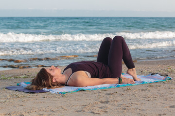 Fototapeta na wymiar Young woman practicing yoga on a mat at the beach. Natural morning light wide shot with the Mediterranean Sea in the background.