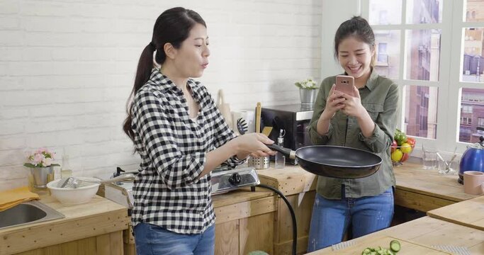 two asian japanese best friends making homemade pancakes in modern kitchen. girl roommate taking photo using smart phone and sharing on social media. happy women successful flipped crepe on pan.