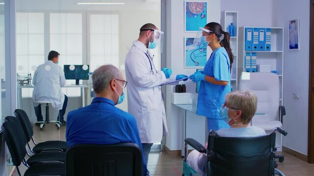 Doctor explaining patient diagnosis to one of nurses wearing protection mask against covid-19 in hospital waiting area. Mature man discussing with handicapped woman in wheelchair.