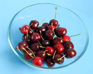 Obraz na płótnie Canvas red ripe cherries in a glass transparent bowl on a blue background side view. summer red berries