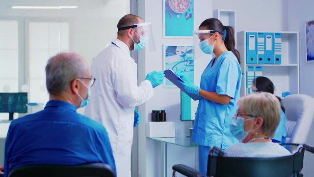 Doctor with face mask and visor against covid-19 giving nurse instruction about patient treatment in hospital waiting area. Disabled senior woman in wheelchair waiting for examination.