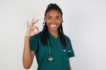 Glad attractive doctor woman shows ok sign with hand as expresses approval, has cheerful expression being optimistic. Standing against gray wall.