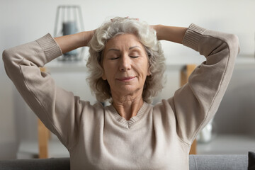 Close up elderly woman puts hands behind head resting in cozy living room enjoy free carefree time, breath fresh conditioning air. Harmony, modern house, healthy pensioner relaxation at home concept