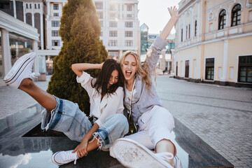 Fototapeta premium Laughing brunette woman having fun with her best friend in spring morning. Carefree sisters in good mood smiling on city background.