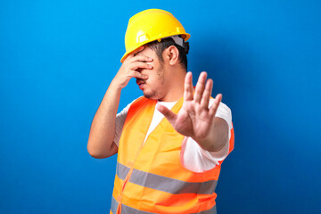 Fat asian construction worker man wearing uniform and helmet over isolated blue background covering...
