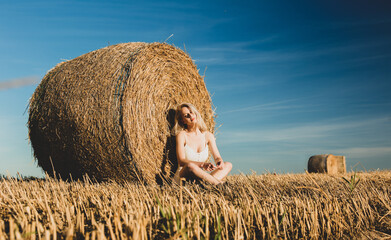 blonde girl sits by a rolled haystack in field in sunset time