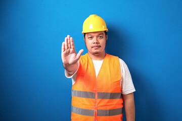 Fat asian workman wearing orange safety vest and yellow helmet making stop gesture