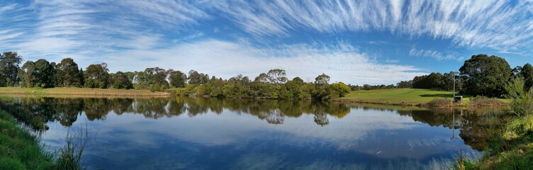 Beautiful afternoon panoramic view of a peaceful pond in a park with reflections of deep blue sky, light clouds and trees on water, Fagan park, Galston, Sydney, New South Wales, Australia