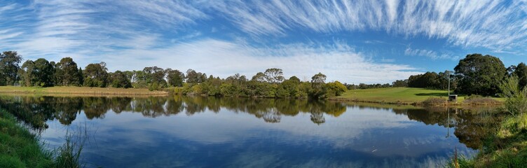 Fototapeta na wymiar Beautiful afternoon panoramic view of a peaceful pond in a park with reflections of deep blue sky, light clouds and trees on water, Fagan park, Galston, Sydney, New South Wales, Australia