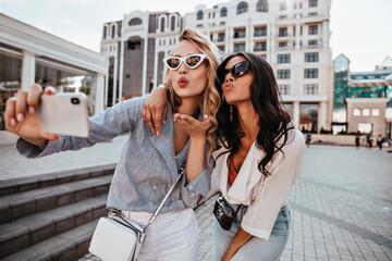 Happy blonde girl in white sunglasses making selfie with kissing face expression. Glamorous female friends expressing love for photo.