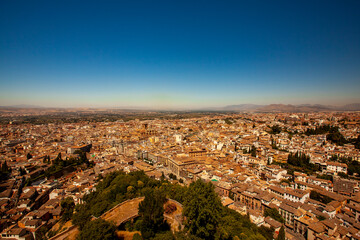 Fototapeta na wymiar Aerial view of the historic Spanish city of Granada featuring rooftops of buildings, cathedrals, roads, trees, naked mountains in background as well as a clear blue sky.
