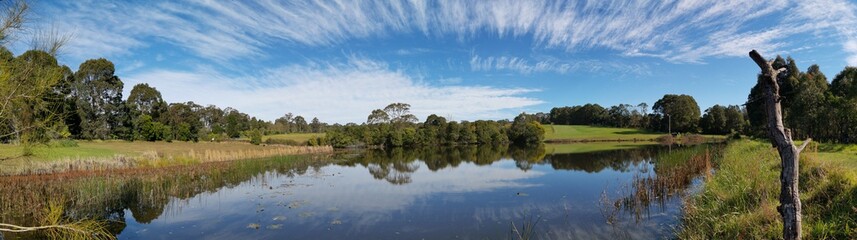 Beautiful afternoon panoramic view of a peaceful pond in a park with reflections of deep blue sky, light clouds and trees on water, Fagan park, Galston, Sydney, New South Wales, Australia