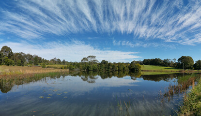 Beautiful afternoon panoramic view of a peaceful pond in a park with reflections of deep blue sky, light clouds and trees on water, Fagan park, Galston, Sydney, New South Wales, Australia
