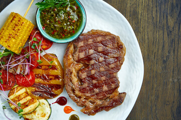 Beef rib eye steak with grilled vegetables and green sauce in a white plate