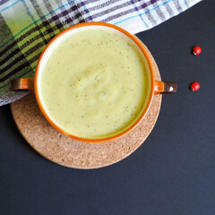 cream soup of broccoli, potatoes, onions and cream in a brown plate with handles on a wooden stand and a white striped towel on a dark background. gluten free food prepared at home