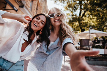 Fascinating blonde girl making selfie on the street with friend. Stunning young ladies enjoying spring day. - obrazy, fototapety, plakaty
