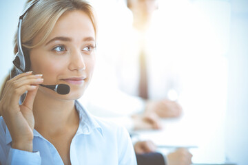 Cheerful smiling business woman with headphones consulting clients. Group of diverse phone operators at work in sunny office.Call center and business people concept