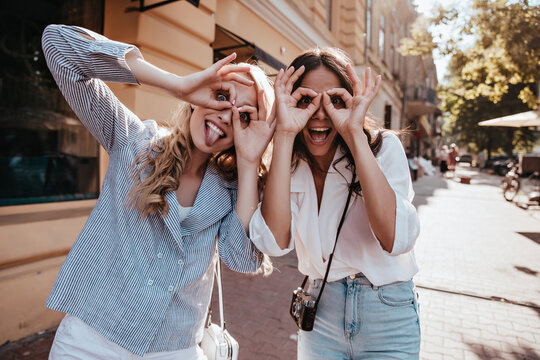 Enchanting young ladies making funny faces on the street. Outdoor photo of elegant female models fooling around together.