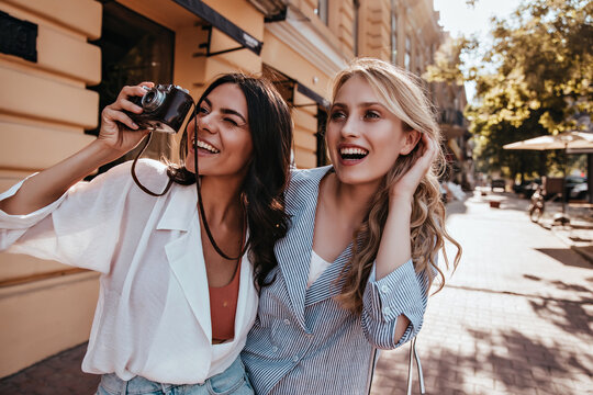 Pretty tanned girl standing on the street with camera. Laughing gorgeous woman taking pictures during walk with friend.
