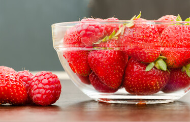 Transparent bowl with strawberries and raspberries close-up side view, berries lie near the bowl.