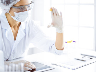 Professional female scientist in protective eyeglasses researching tube with reagents in...