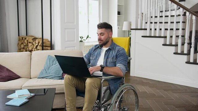 Handsome smiling friendly modern young bearded guy in wheelchair having video call on laptop with relatives or friends.being at home