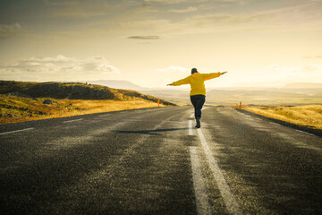 Iceland road trip. Girl is walking alone on road to sunset and keeping balance on dividing strip line. Freedom concept. Beautiful icelandic landscape