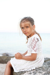 Fototapeta na wymiar A small and pretty girl with blond hair and blue eyes, sits on the beach, on large stones by the sea. Summer Holidays Vacations.