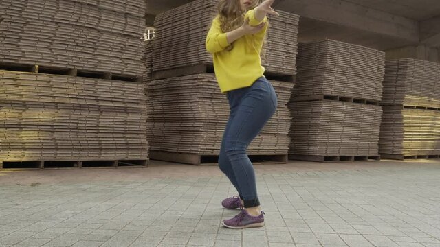 long shot 4K. modern choreography concept. A young plastic girl in a bright yellow sweater contemporary dancer is dancing on the street on an industrial background.