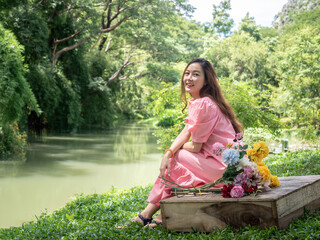 Asian woman in pink dress with a bouquet of flowers sitting in the garden near canal.