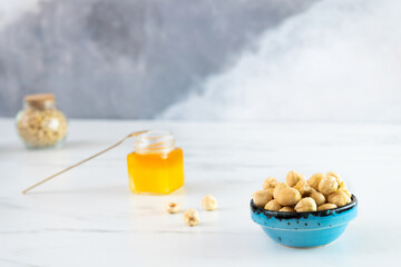 Peeled roasted hazelnuts in a blue bowl stands on a white plate. Nearby there is a small glass jar with honey and a spoon, and there are several nuts on the table. Delicious and healthy sweets.