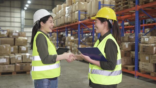 wholesale logistic people and export concept. two female manual workers  with clipboard and tablet shaking hands and making deal at warehouse. asian women employees start up teamwork in stockroom.