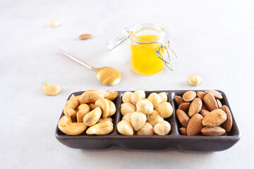 Various peeled nuts (cashews, hazelnuts, almonds) in a black bowl with three branches stand against a gray concrete background. There is a jar of honey and a small spoon next to it. Healthy dessert