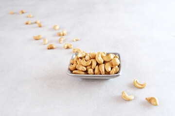 Fototapeta na wymiar A gray rectangular bowl of peeled roasted cashews stands on a light gray concrete background. Cashew nuts lie in one diagonal line. Healthy food high in antioxidants, protein and vitamins. Top view
