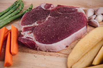 Raw meat t-bone steak with herbs, spices, potato, carrot, garlic and asparagus on a wood background