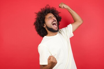 Attractive young caucasian man celebrating a victory punching the air with his fists and a beaming toothy smile over a blue studio background with copy space. - obrazy, fototapety, plakaty