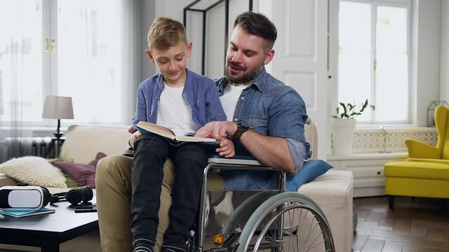 Family concept of good-tempered lucky young bearded disabled dad in wheelchair which helping to read book to his small son that sitting on his knees