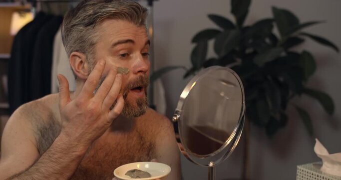 Mature good looking man applying facial mask while sitting in front of mirror. Bearded guy in 40s doing beauty prosedure in evening at home. Concept of care and beauty.