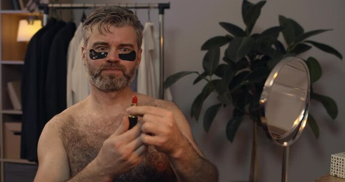 Brutal mature bachelor opening and closing red lipstick while shaking head. Bearded guy in 40s with eye patches sitting in front of mirror at home in evening. Concept of beauty.