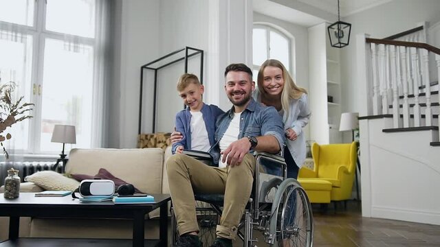 Close up of good-natured smiling lucky young mother,father in wheelchair and their son which posing on camera in beautifully designeted apartment