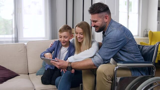 Good-looking happy smiling modern bearded dad in wheelchair,mother and son using i-pad in contemporary living-room