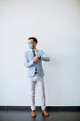 Young and stylish business man with face mask stands by the wall in the office and checking time