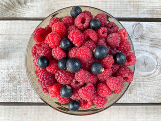 Ripe raspberries and blackcurrants in a glass bowl on a wooden table. The photo at the top. Harvest. Fresh summer berries.