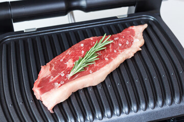 A piece of fresh meat with coarse salt and a sprig of rosemary. Juicy beef steak on the electric grill. - 368849972