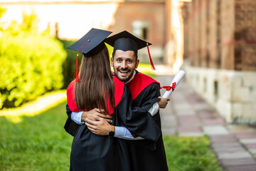 Graduates in mantles with diplomas in hands are standing near university and hugging each other.