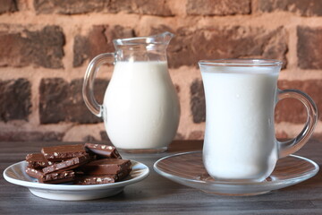 milk in a transparent cup and jug and chocolate wedges on a transparent saucer against a brick wall background